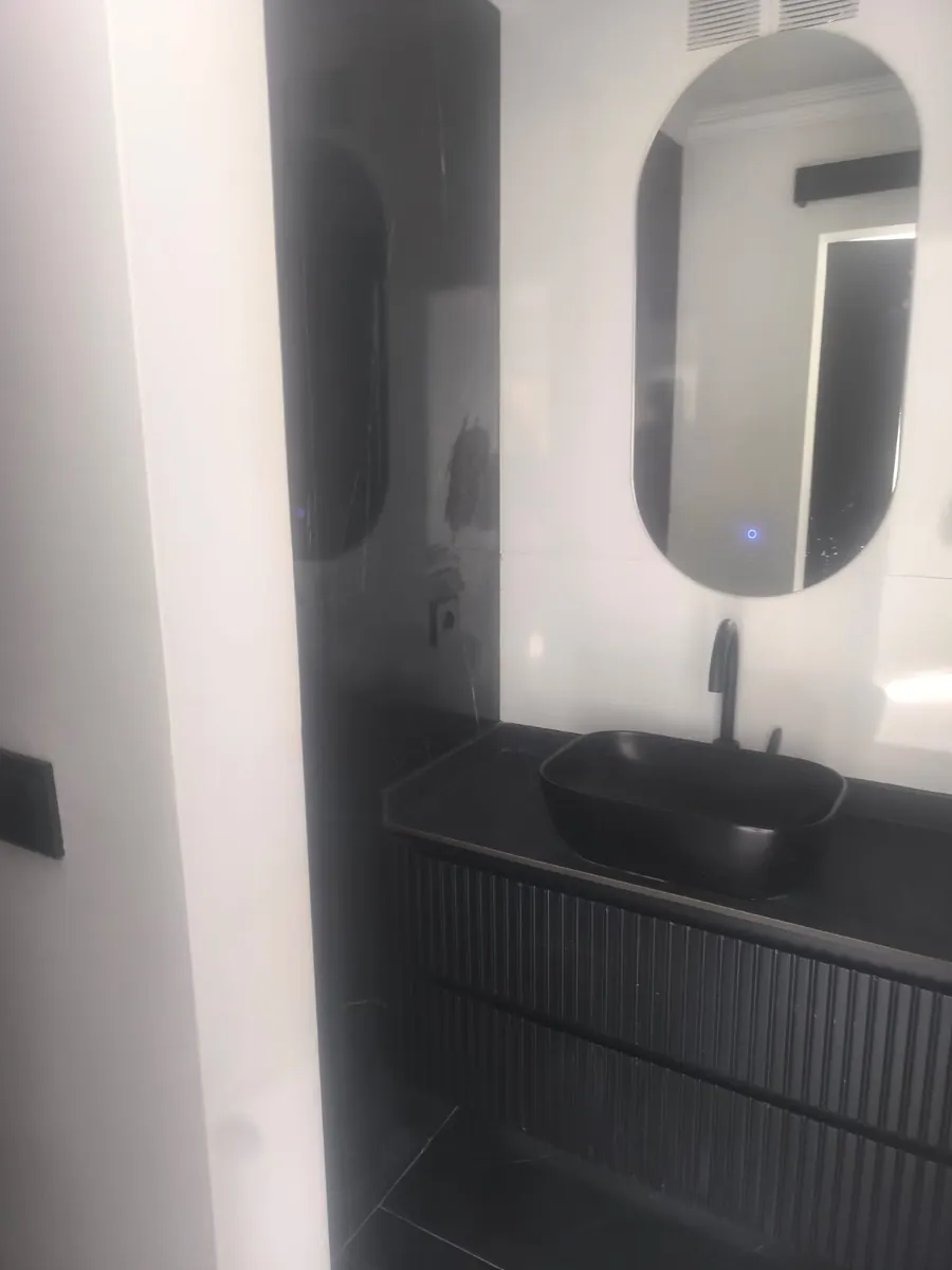 Finished bathroom vanity with a matte black basin, ribbed black cabinet front, black tap and an oval backlit mirror against dark marble-effect tiles